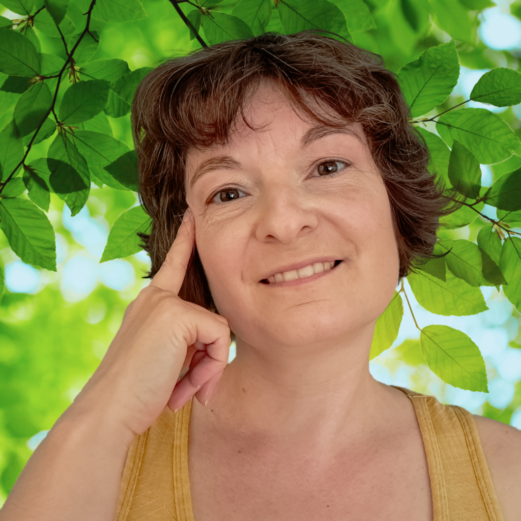 Porträtfoto Lesley vor einem Blätterhintergrund mit Finger denkend, fragend an der Schläfe